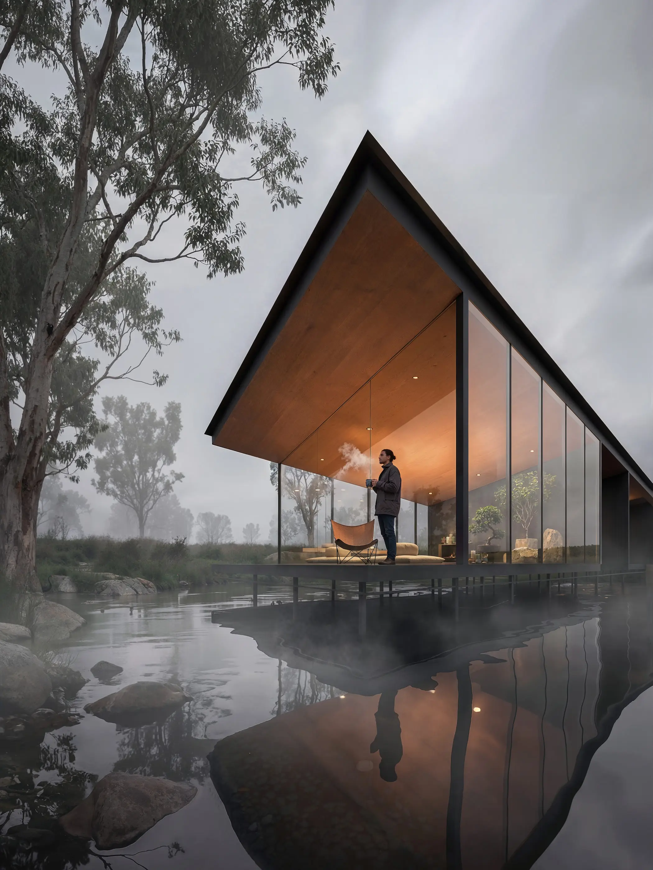 The gabled wellness pavilion at Beaconsfield stands on slender steel columns above the dam in heavy fog, its timber-lined ceiling lit warm behind glass walls, gum trees and boulders beyond.