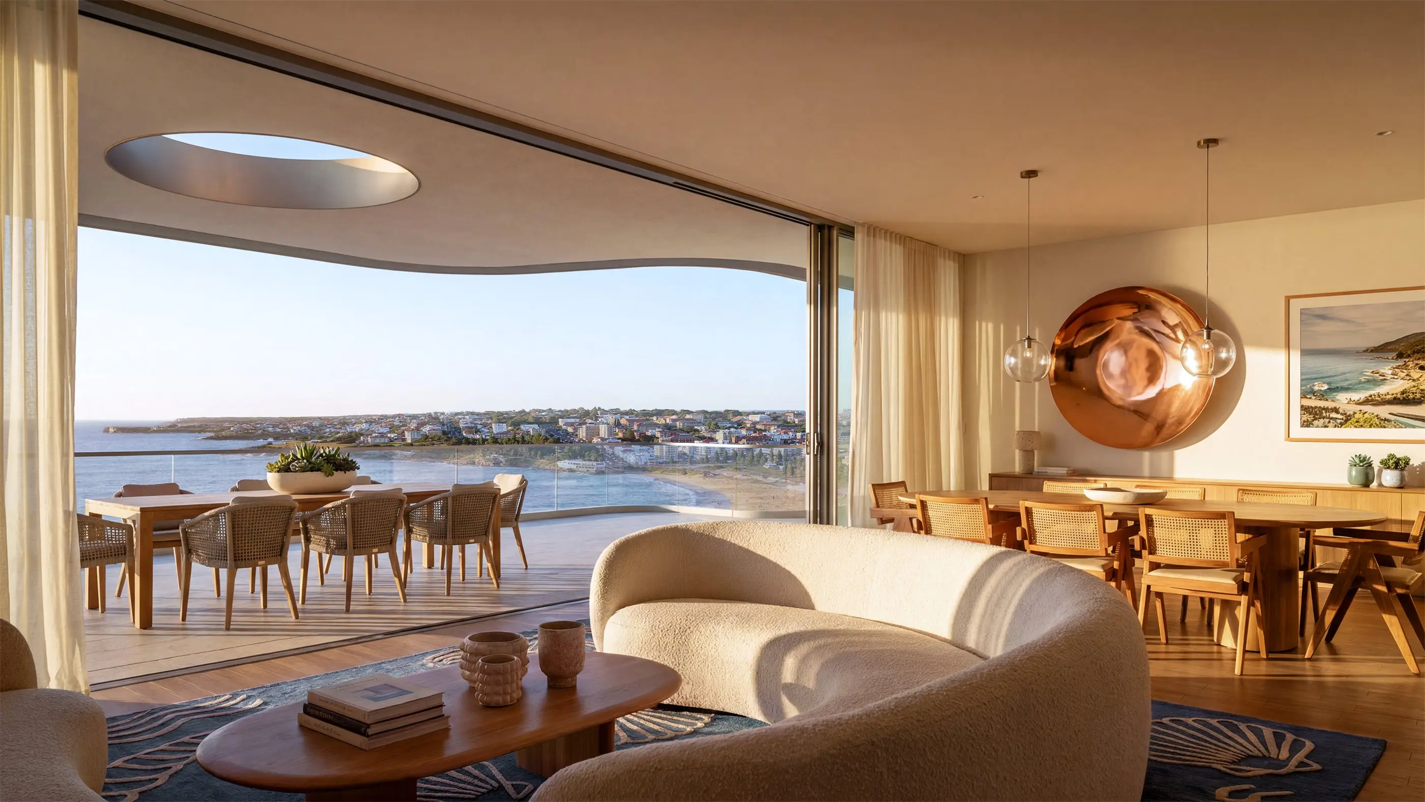 Living and dining room in a Coogee apartment with a curved ceiling oculus, a copper wall disc, and glazing sliding open to a terrace with a long outdoor table and views across the bay to the headland